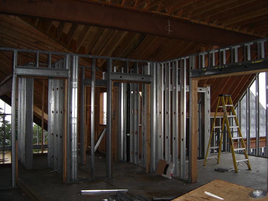 Wnętrze budynku w trakcie budowy, widoczne metalowe profile ścian działowych, drewniany strop i żółta drabina.